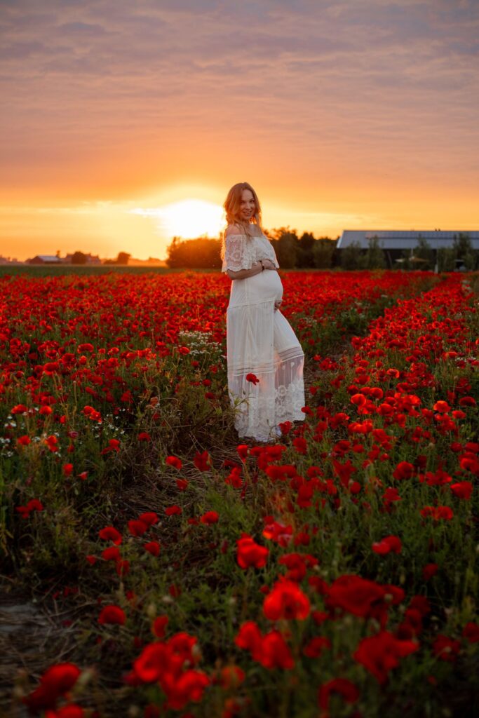 Zwangere vrouw in creme jurk tijdens zonsondergang met 1 hand op en 1 hand onder buik