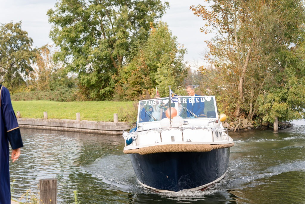 Bruidspaar komt aanvaren met versierde boot ‘Just Married’ De boot met het bord ‘Just Married’ nadert de aanlegsteiger, met het bruidspaar dat vrolijk naar de gasten zwaait.