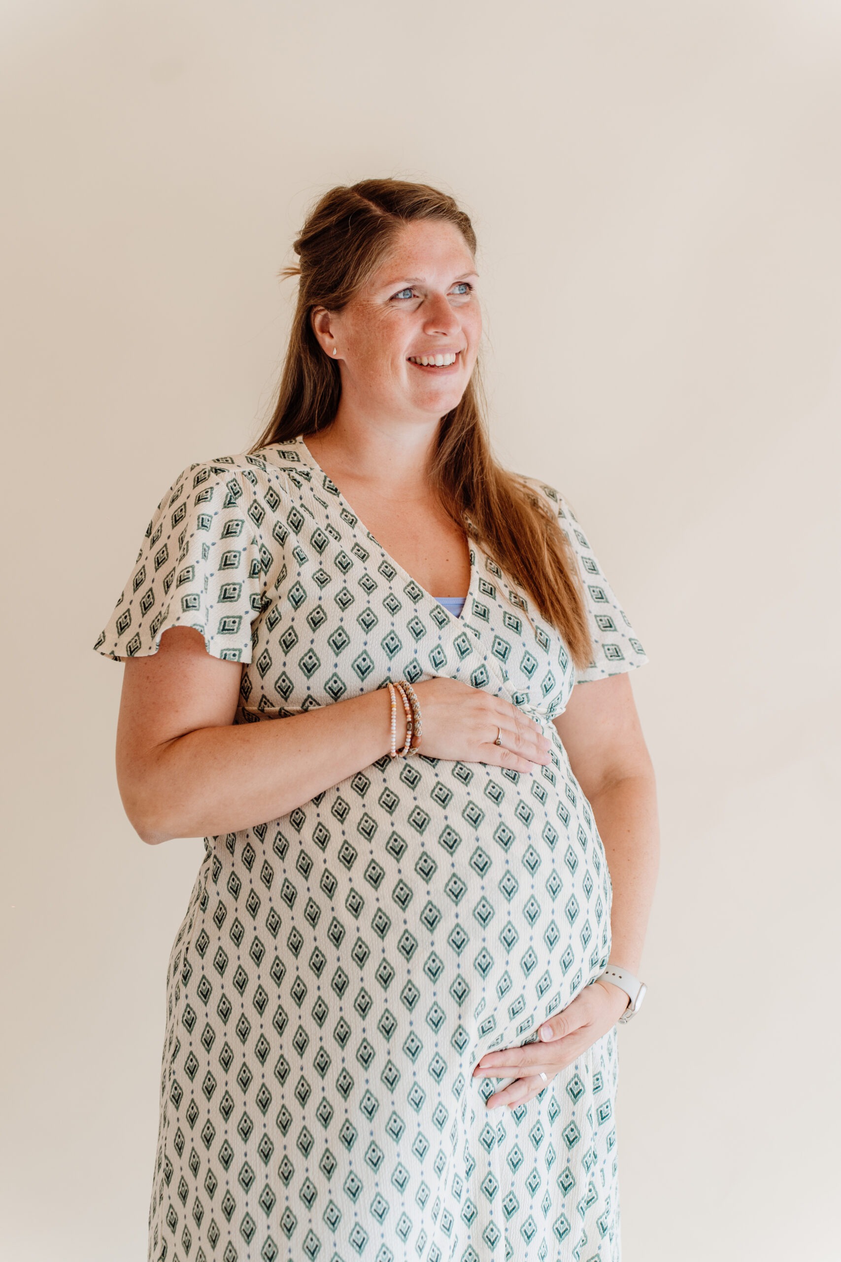 Zwangere vrouw lacht terwijl ze met haar handen haar buik vasthoudt, lichte achtergrond, zachte belichting.