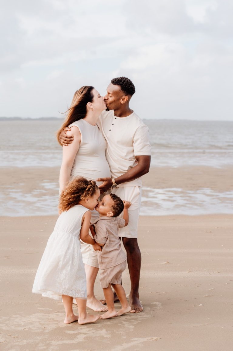 Papa bukt zich naar hun kind toe terwijl Daisy zwanger naast hen staat, zachte gouden tinten op het strand
