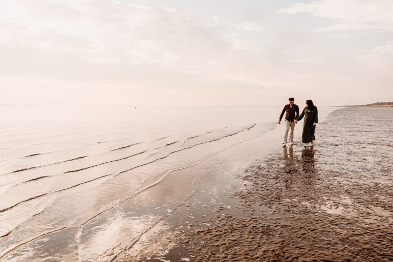 Zwangere vrouw en partner lopen hand in hand langs de vloedlijn op het brede strand van Rockanje, wijd panoramisch beeld