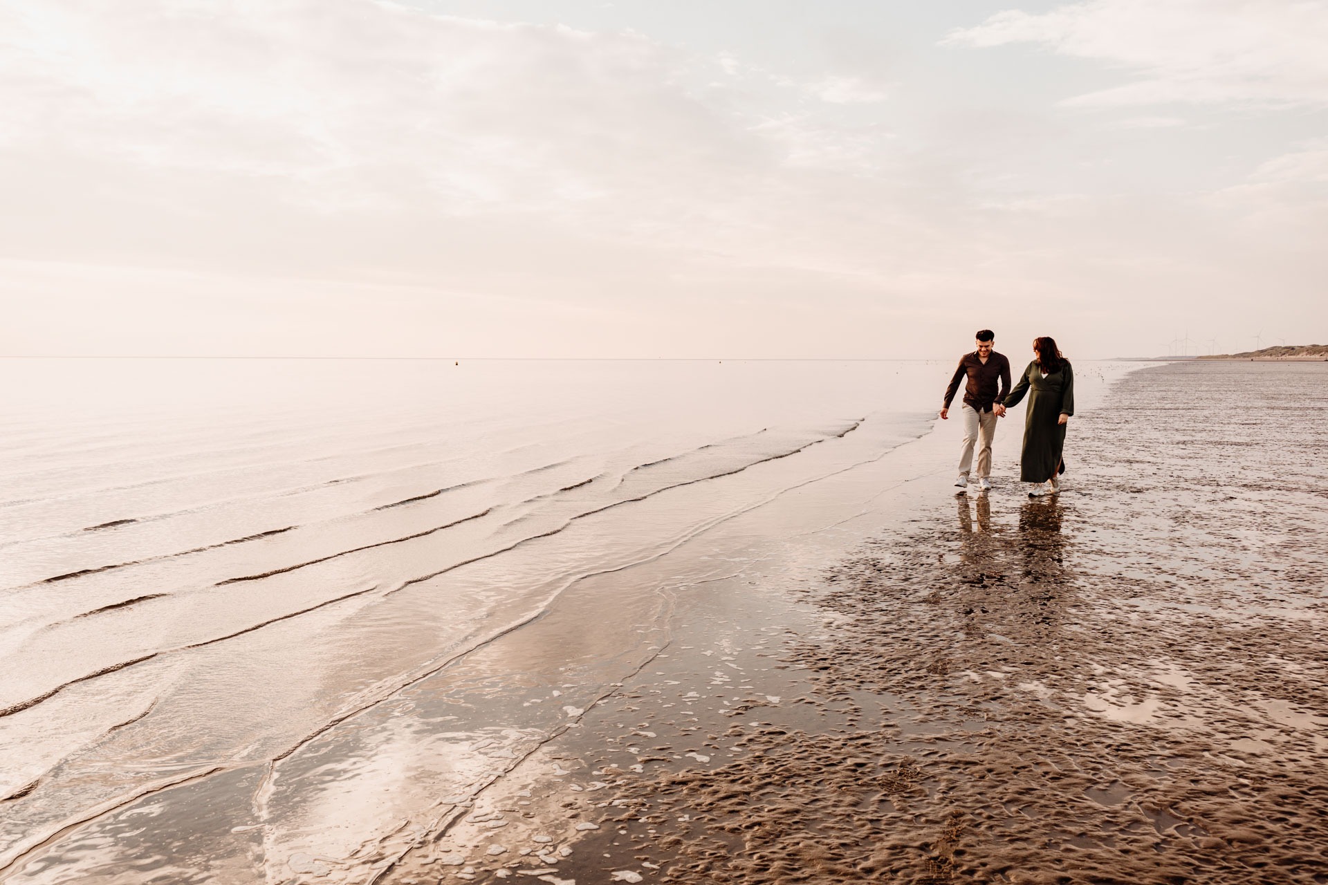 Zwangere vrouw en partner lopen hand in hand langs de vloedlijn op het brede strand van Rockanje, wijd panoramisch beeld